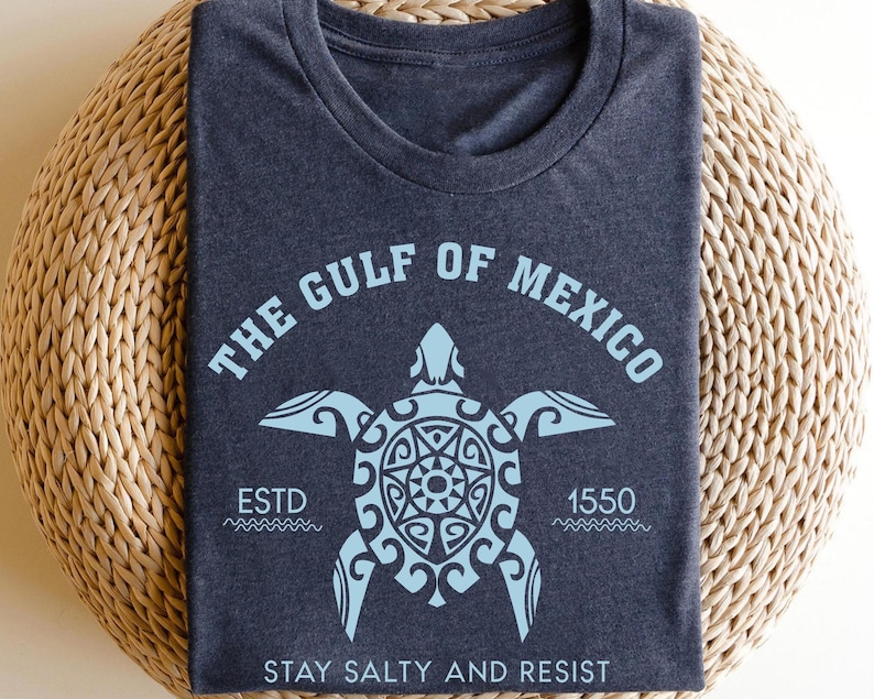 May include: Dark blue t-shirt with a light blue graphic of a sea turtle and the text "THE GULF OF MEXICO". Below the turtle are the words "ESTD 1550" and "STAY SALTY AND RESIST". The shirt is folded on a woven surface.