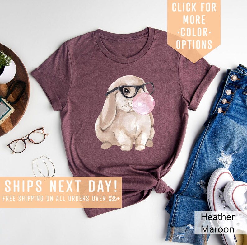May include: A heather maroon t-shirt with a watercolor illustration of a bunny wearing glasses and blowing a bubble. The text "SHIPS NEXT DAY! FREE SHIPPING ON ALL ORDERS OVER $35+" is at the bottom of the image.