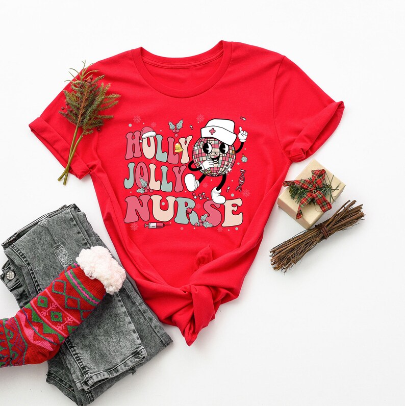Op de afbeelding: Een rood t-shirt met de woorden "HOLLY JOLLY NURSE" in kleurrijke retro letters. Het shirt heeft een cartoon discobal karakter met een verpleegstershoed. Het shirt wordt gecombineerd met een grijze jeans en een kerstsok.