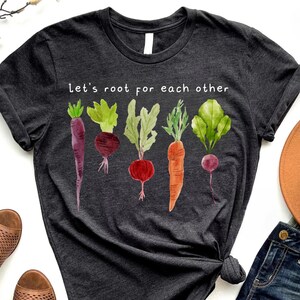 May include: Dark gray t-shirt with a watercolor illustration of various root vegetables. The shirt features the text "Let's root for each other" in white, promoting a gardening or healthy eating theme.