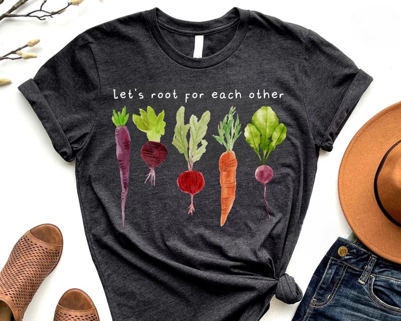 May include: Dark gray t-shirt with a watercolor illustration of various root vegetables. The shirt features the text "Let's root for each other" in white, promoting a gardening or healthy eating theme.