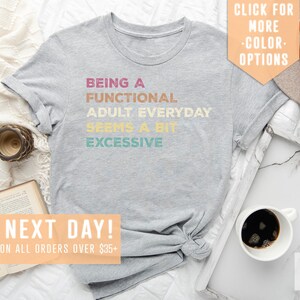 May include: Gray t-shirt with the text "Being a Functional Adult Everyday Seems a Bit Excessive" in a rainbow color scheme. The shirt is laid flat on a white surface with a book, a clock, a cup of coffee, and a hat.