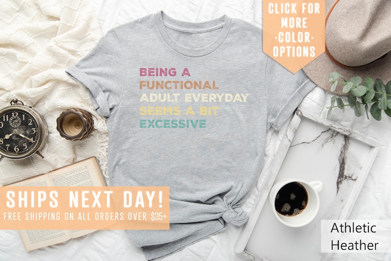 May include: Gray t-shirt with the text "Being a Functional Adult Everyday Seems a Bit Excessive" in a rainbow color scheme. The shirt is laid flat on a white surface with a book, a clock, a cup of coffee, and a hat.
