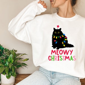 May include: White sweatshirt with a black cat wearing Christmas lights and a red star on its head. The text "MEOWY CHRISTMAS" is printed below the cat in rainbow colors.