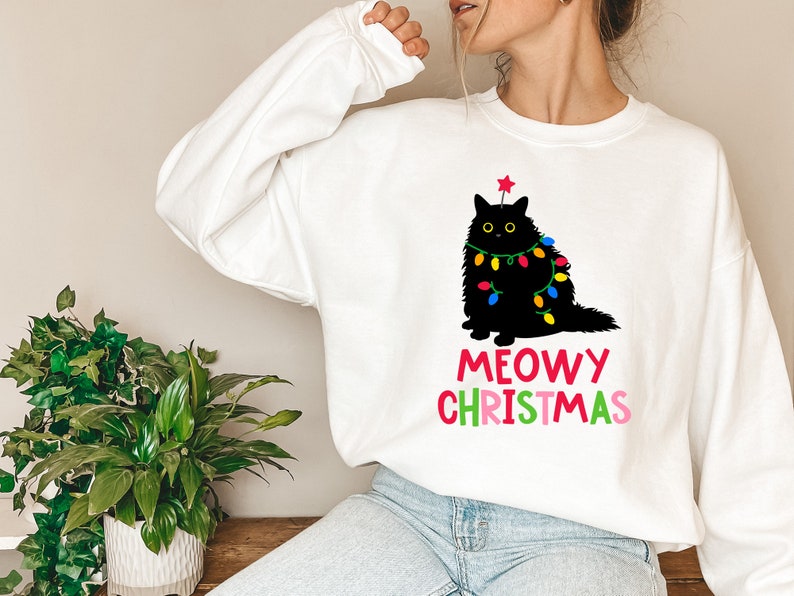 May include: White sweatshirt with a black cat wearing Christmas lights and a red star on its head. The text "MEOWY CHRISTMAS" is printed below the cat in rainbow colors.