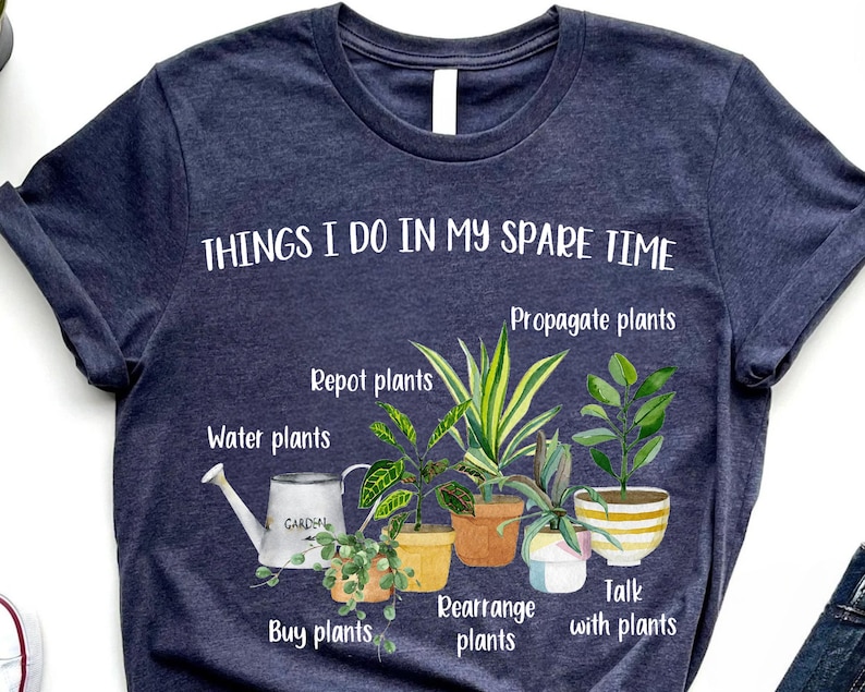 以下が含まれることがあります： 「THINGS I DO IN MY SPARE TIME」というテキストと、植物に関連する活動のリスト（「Propagate plants」、「Repot plants」、「Water plants」、「Buy plants」、「Rearrange plants」、「Talk with plants」など）が書かれたネイビーブルーのTシャツ。テキストは白で、画像には鉢植えの植物のイラストが含まれています。