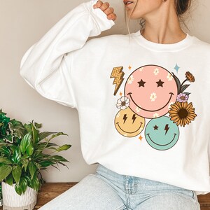 May include: White sweatshirt with a graphic design of three smiley faces with flowers and lightning bolts. The smiley faces are pink, yellow, and green.