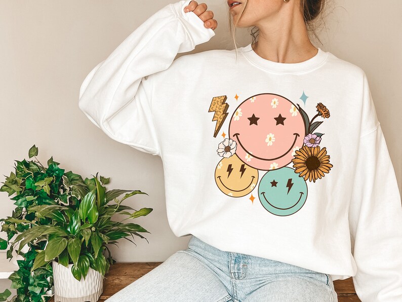 May include: White sweatshirt with a graphic design of three smiley faces with flowers and lightning bolts. The smiley faces are pink, yellow, and green.