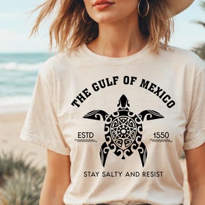 May include: Cream-colored t-shirt featuring a black turtle design with the text "THE GULF OF MEXICO" and "ESTD 1550". The shirt also includes the phrase "STAY SALTY AND RESIST".