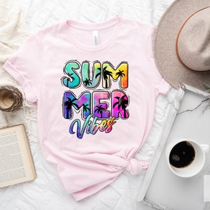 May include: Pink t-shirt with the words "SUMMER VIBES" in a colorful, tropical design. The text features palm tree silhouettes and a tie-dye effect, evoking a summer theme. The shirt is displayed on a white surface.