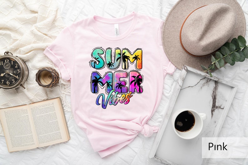 May include: Pink t-shirt with the words "SUMMER VIBES" in a colorful, tropical design. The text features palm tree silhouettes and a tie-dye effect, evoking a summer theme. The shirt is displayed on a white surface.