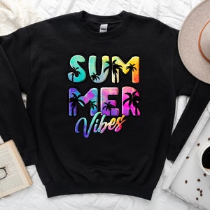 May include: Black sweatshirt with the words "SUMMER VIBES" in colorful, tropical-themed lettering. The design includes palm tree silhouettes. A hat, book, and coffee cup are in the background.