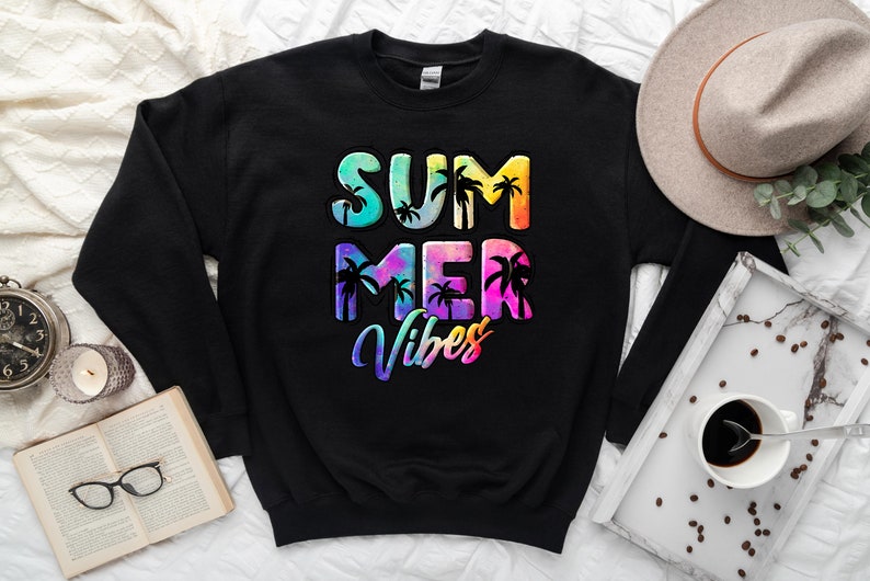May include: Black sweatshirt with the words "SUMMER VIBES" in colorful, tropical-themed lettering. The design includes palm tree silhouettes. A hat, book, and coffee cup are in the background.