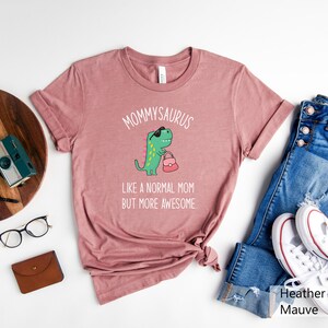 May include: Heather mauve t-shirt with the text "MOMMYSAURUS" above a cartoon dinosaur wearing sunglasses and holding a purse. The text "LIKE A NORMAL MOM BUT MORE AWESOME" is below the dinosaur. The shirt is paired with blue jeans and white sneakers.