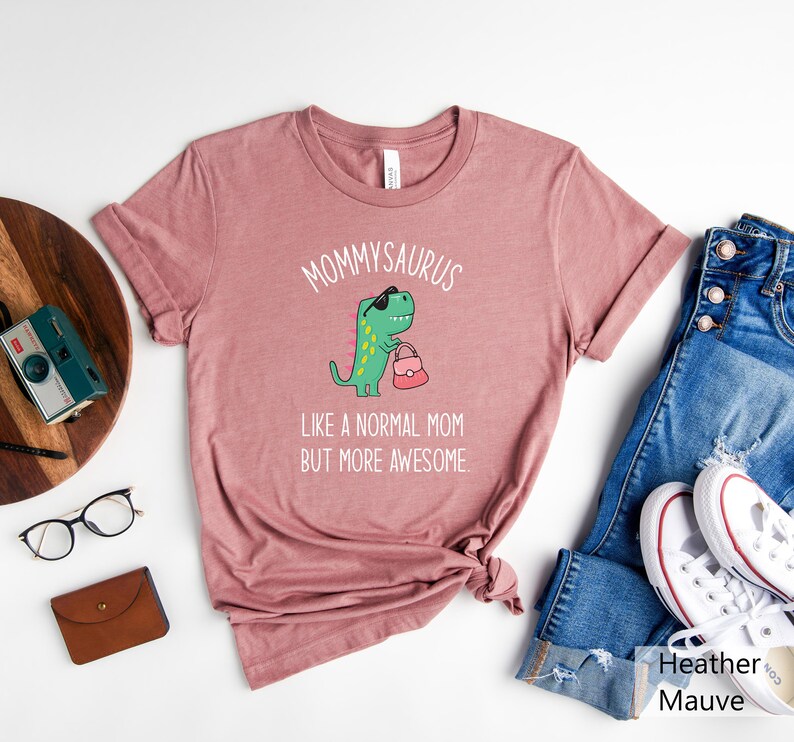 May include: Heather mauve t-shirt with the text "MOMMYSAURUS" above a cartoon dinosaur wearing sunglasses and holding a purse. The text "LIKE A NORMAL MOM BUT MORE AWESOME" is below the dinosaur. The shirt is paired with blue jeans and white sneakers.