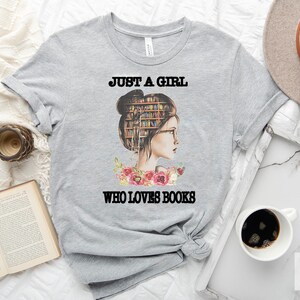 Pu&ograve; includere: Maglietta grigia con una grafica di una donna con libri nei capelli e il testo "Just a girl who loves books".