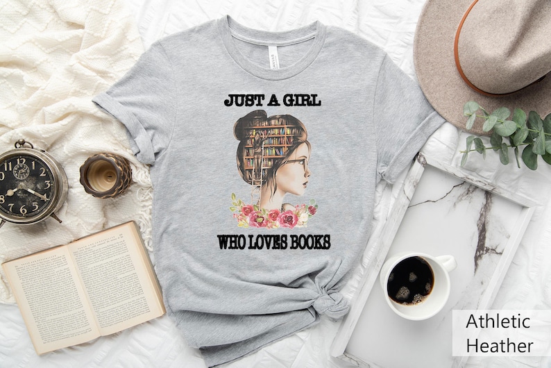 Pu&ograve; includere: Maglietta grigia con una grafica di una donna con libri nei capelli e il testo "Just a girl who loves books".