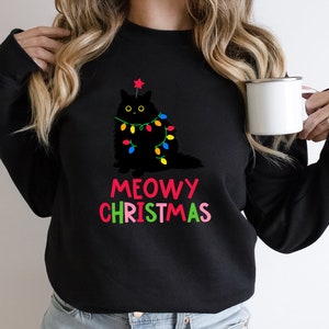 May include: Black sweatshirt with a black cat wearing Christmas lights and a star on its head. The text "MEOWY CHRISTMAS" is written in colorful letters below the cat.