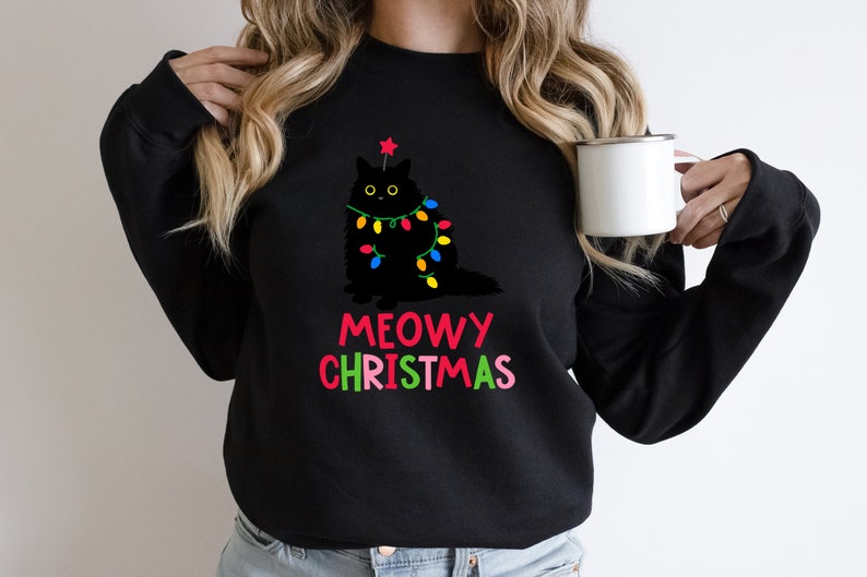 May include: Black sweatshirt with a black cat wearing Christmas lights and a star on its head. The text "MEOWY CHRISTMAS" is written in colorful letters below the cat.