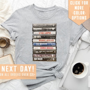 May include: A heather grey t-shirt with a graphic of a stack of vintage cassette tapes featuring artists like Madonna, Cyndi Lauper, and Van Halen. The text "SHIPS NEXT DAY! FREE SHIPPING ON ALL ORDERS OVER $35+" is at the bottom of the image.