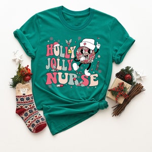 Op de afbeelding: Petrolkleurig T-shirt met de tekst "Holly Jolly Nurse" in een retro lettertype, met een discobal-personage met een verpleegsterspet. Het ontwerp bevat sneeuwvlokken, hulst en een spuit. Kerst-thema kleding.