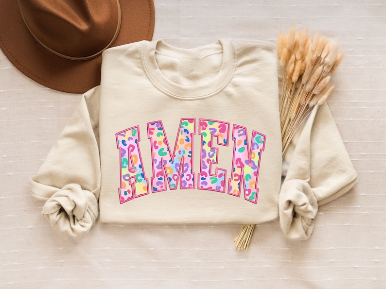 Puede incluir: Una sudadera beige con la palabra "AMEN" impresa en letras grandes y coloridas. Las letras est&aacute;n rellenas con un estampado de leopardo en tonos de rosa, morado, azul, verde y amarillo.