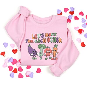 May include: Pink sweatshirt with a graphic of smiling vegetables and the text "Let's Root For Each Other".