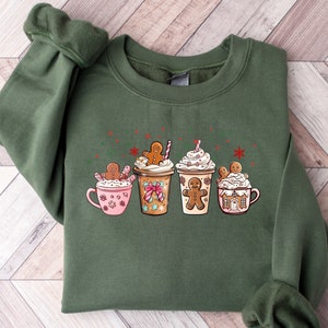 Puede incluir: Sudadera verde con un dise&ntilde;o navide&ntilde;o con cuatro tazas con hombres de jengibre y crema batida. Las tazas est&aacute;n llenas de diferentes bebidas, incluyendo chocolate caliente, caf&eacute; y un latte de jengibre.