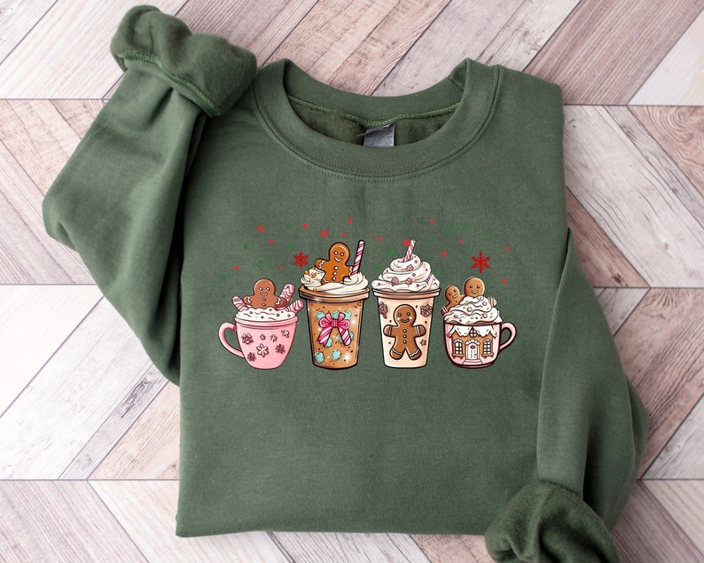 Puede incluir: Sudadera verde con un dise&ntilde;o navide&ntilde;o con cuatro tazas con hombres de jengibre y crema batida. Las tazas est&aacute;n llenas de diferentes bebidas, incluyendo chocolate caliente, caf&eacute; y un latte de jengibre.