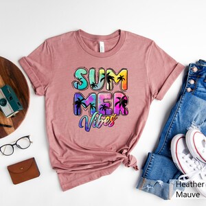 May include: Heather mauve t-shirt with the words "SUMMER VIBES" in a colorful, tropical design. The text features palm tree silhouettes within the letters. The shirt is displayed with blue jeans and white sneakers.