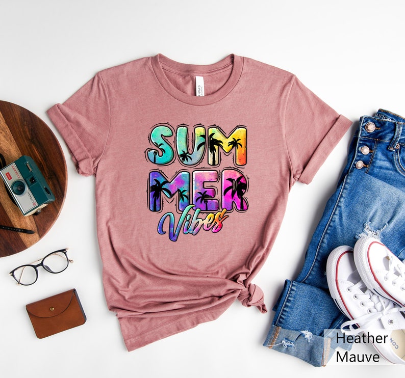 May include: Heather mauve t-shirt with the words "SUMMER VIBES" in a colorful, tropical design. The text features palm tree silhouettes within the letters. The shirt is displayed with blue jeans and white sneakers.