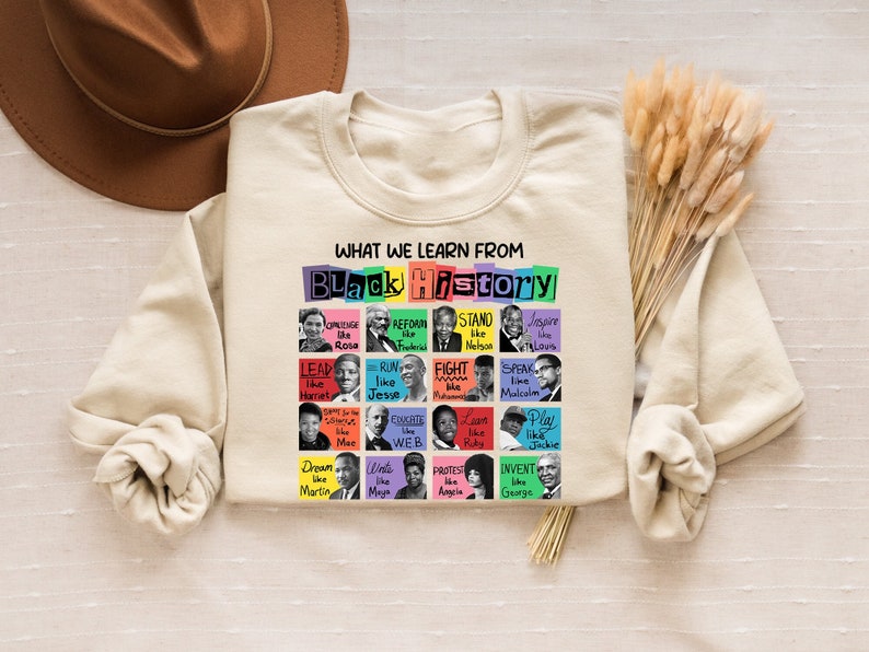 May include: A beige sweatshirt with the text 'WHAT WE LEARN FROM BLACK HISTORY' in black letters. The text is surrounded by a colorful grid of squares with images of notable Black figures and phrases like 'LEAD like Harriet', 'RUN like Jesse', 'FIGHT like Muhammad', 'SPEAK like Malcolm', 'COACH like WEB', 'PLAY like Jackie', 'DREAM like Martin', 'WRITE like Maya', 'PROTEST like Angela', 'INVENT like George'.