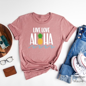 May include: Heather mauve t-shirt with the words "LIVE. LOVE. ALOHA" in white, a yellow pineapple, and blue waves. The shirt is displayed with blue jeans and white sneakers.