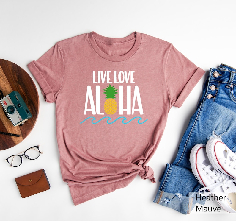 May include: Heather mauve t-shirt with the words "LIVE. LOVE. ALOHA" in white, a yellow pineapple, and blue waves. The shirt is displayed with blue jeans and white sneakers.