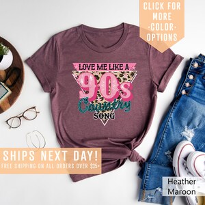 May include: A maroon t-shirt featuring a pink and white graphic that reads "Love Me Like A 90's Country Song". The graphic is in a triangular shape with a leopard print background.