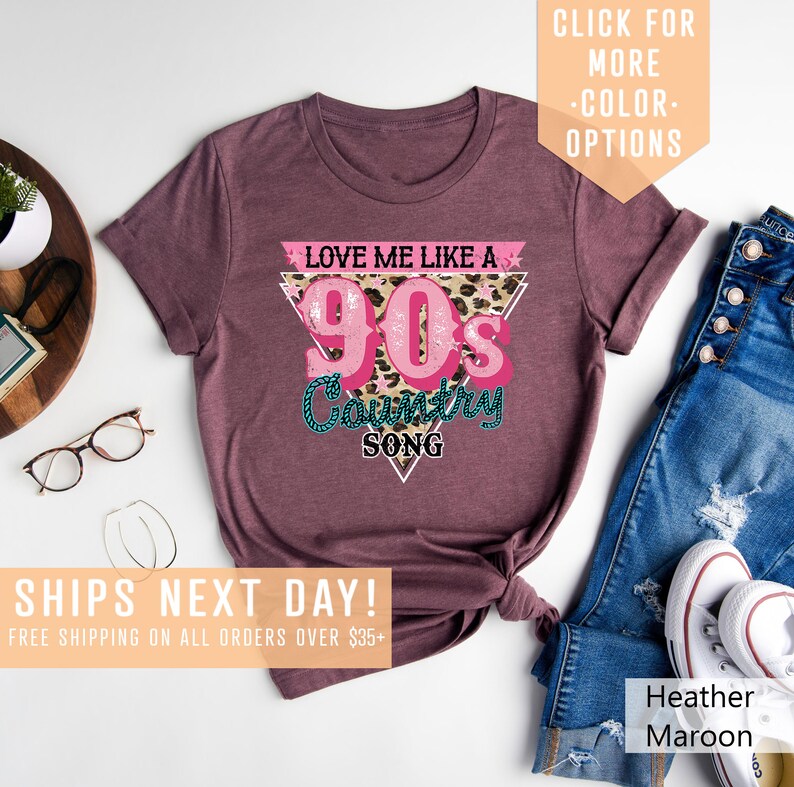 May include: A maroon t-shirt featuring a pink and white graphic that reads "Love Me Like A 90's Country Song". The graphic is in a triangular shape with a leopard print background.