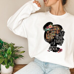 May include: White sweatshirt with a graphic of a woman's silhouette with a bookshelf for a head. The text reads "& into the library she goes to lose her MIND and find her SOUL."