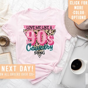 May include: Pink t-shirt featuring a graphic design that reads "Love Me Like A 90s Country Song" in a pink and teal colour scheme with a leopard print background.
