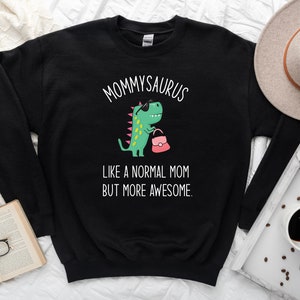 May include: Black sweatshirt with the text "Mommysaurus" above a cartoon dinosaur wearing sunglasses and holding a pink purse. The text "Like a normal mom but more awesome" is below the dinosaur.