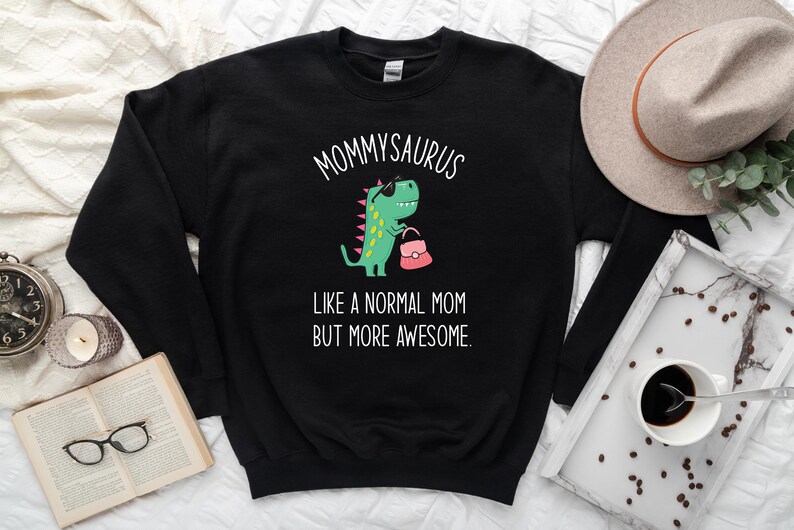May include: Black sweatshirt with the text "Mommysaurus" above a cartoon dinosaur wearing sunglasses and holding a pink purse. The text "Like a normal mom but more awesome" is below the dinosaur.