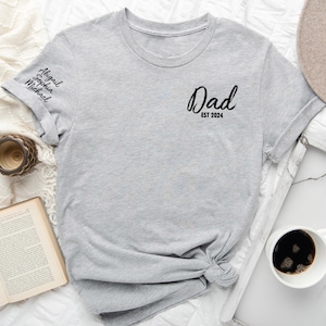 May include: Gray t-shirt with the text "Dad Est 2024" on the front and the names "Abigail, Sophia, Michael" on the sleeve.