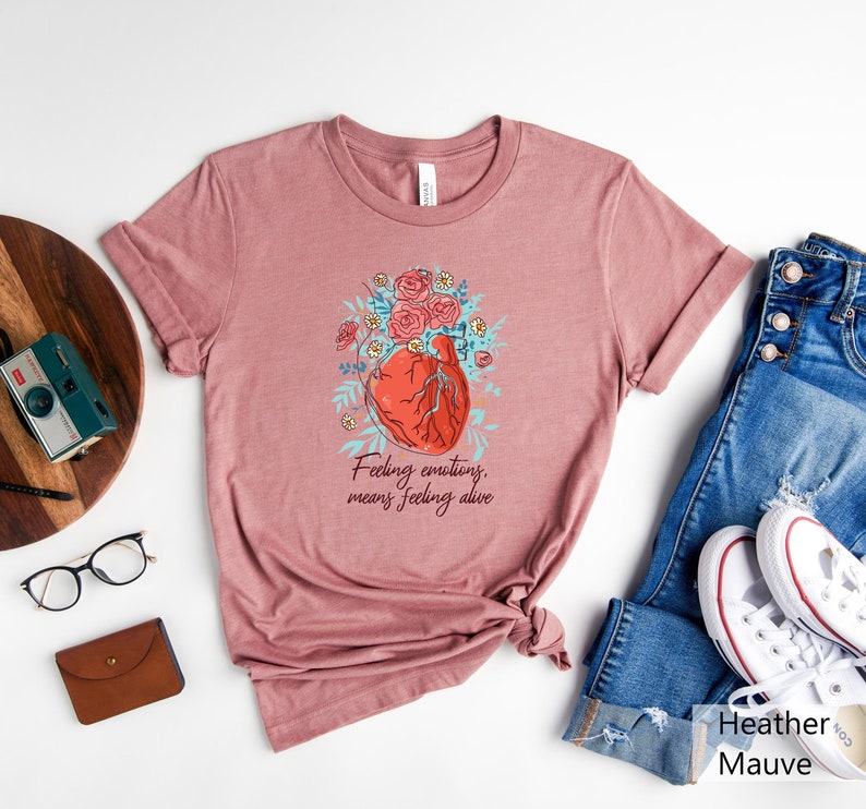 May include: A heather mauve t-shirt featuring a graphic of a heart with flowers and the text "Feeling emotions, means feeling alive".