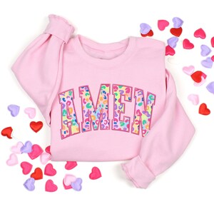 Puede incluir: Sudadera rosa con la palabra "AMEN" en letras de estampado de leopardo arco&iacute;ris.