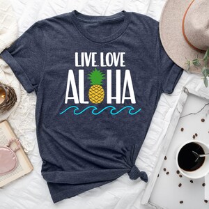 May include: Heather navy t-shirt with the words "LIVE. LOVE. ALOHA" in white, a yellow pineapple, and blue waves. The shirt is knotted at the bottom. A hat, coffee, and a candle are in the background.