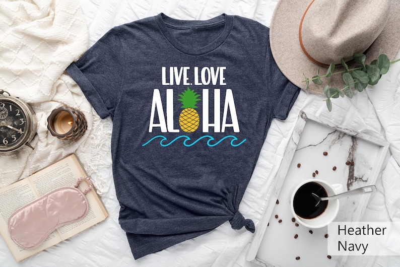 May include: Heather navy t-shirt with the words "LIVE. LOVE. ALOHA" in white, a yellow pineapple, and blue waves. The shirt is knotted at the bottom. A hat, coffee, and a candle are in the background.