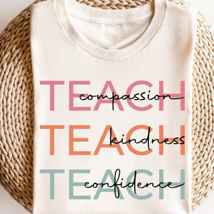 Puede incluir: Una camiseta blanca con las palabras "TEACH compassion", "TEACH kindness" y "TEACH confidence" impresas en diferentes colores.
