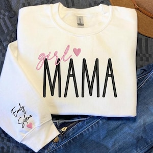Puede incluir: Sudadera blanca con las palabras "girl MAMA" en letras rosas y negras, con un corazón rosa. La manga izquierda tiene los nombres "Emily Selena" y un corazón rosa. La sudadera se combina con vaqueros azules.