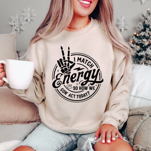 Op de afbeelding: Een beige sweatshirt met een zwarte grafische print van een hand die een peace-teken maakt met de tekst "I Match Energy So How We Gon' Act Today?"