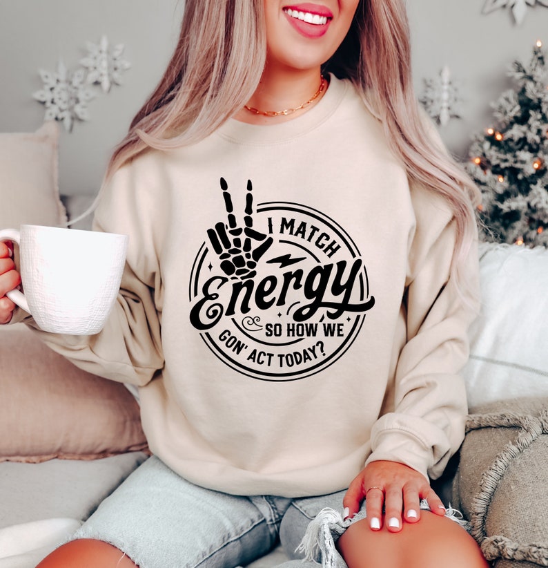 Op de afbeelding: Een beige sweatshirt met een zwarte grafische print van een hand die een peace-teken maakt met de tekst "I Match Energy So How We Gon' Act Today?"