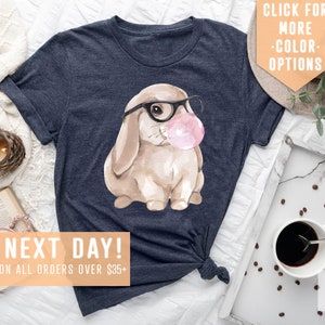 May include: A navy blue t-shirt with a watercolor illustration of a brown bunny wearing glasses and blowing a pink bubble. The text "SHIPS NEXT DAY! FREE SHIPPING ON ALL ORDERS OVER $35+" is at the bottom of the image.